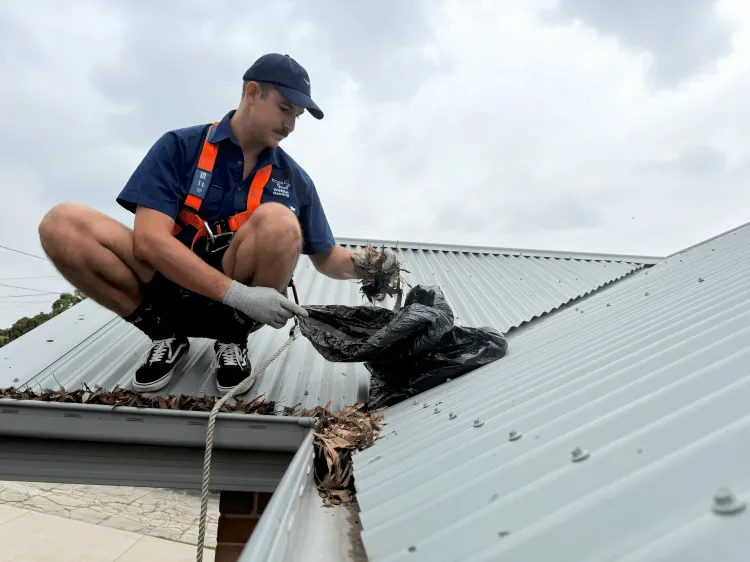 Man in safety harness smiling while cleaning a roof gutter filled with leaves.