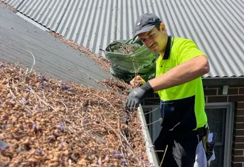 Man in high-vis shirt and gloves cleaning a roof gutter overflowing with dried leaves.